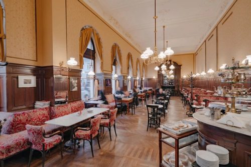 Langer Saal des Café Sperl mit Rundbogenfenstern, Messinglustern, roten Polsterbänken und Marmortischen