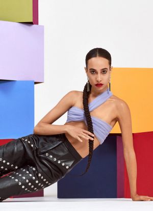A woman with a long braided ponytail wearing black leather pants and a lavender-colored top poses in front of stacks of brightly-colored cubes.