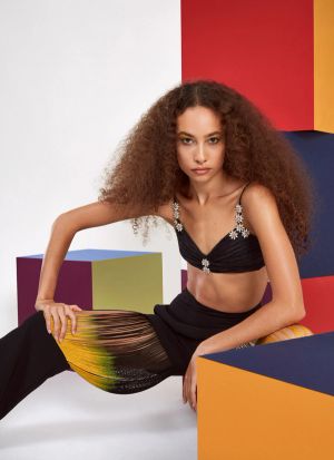 A woman with long curly hair, a black crop-top, and baggy pants crouching among stacks of colorful cubes