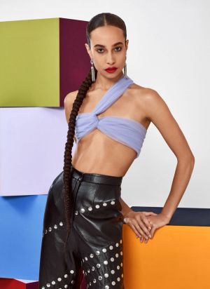 A woman with a long braided ponytail wearing black leather pants and a lavender-colored top standing in front of stacks of brightly-colored cubes.