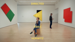 Two people in a gallery of modern art overlayed with the text "DIRECTED BY EZRA HURWITZ"