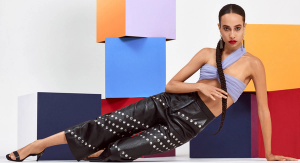 A woman with a long braided ponytail wearing black leather pants and a lavender-colored top posing sideways in front of stacks of brightly-colored cubes