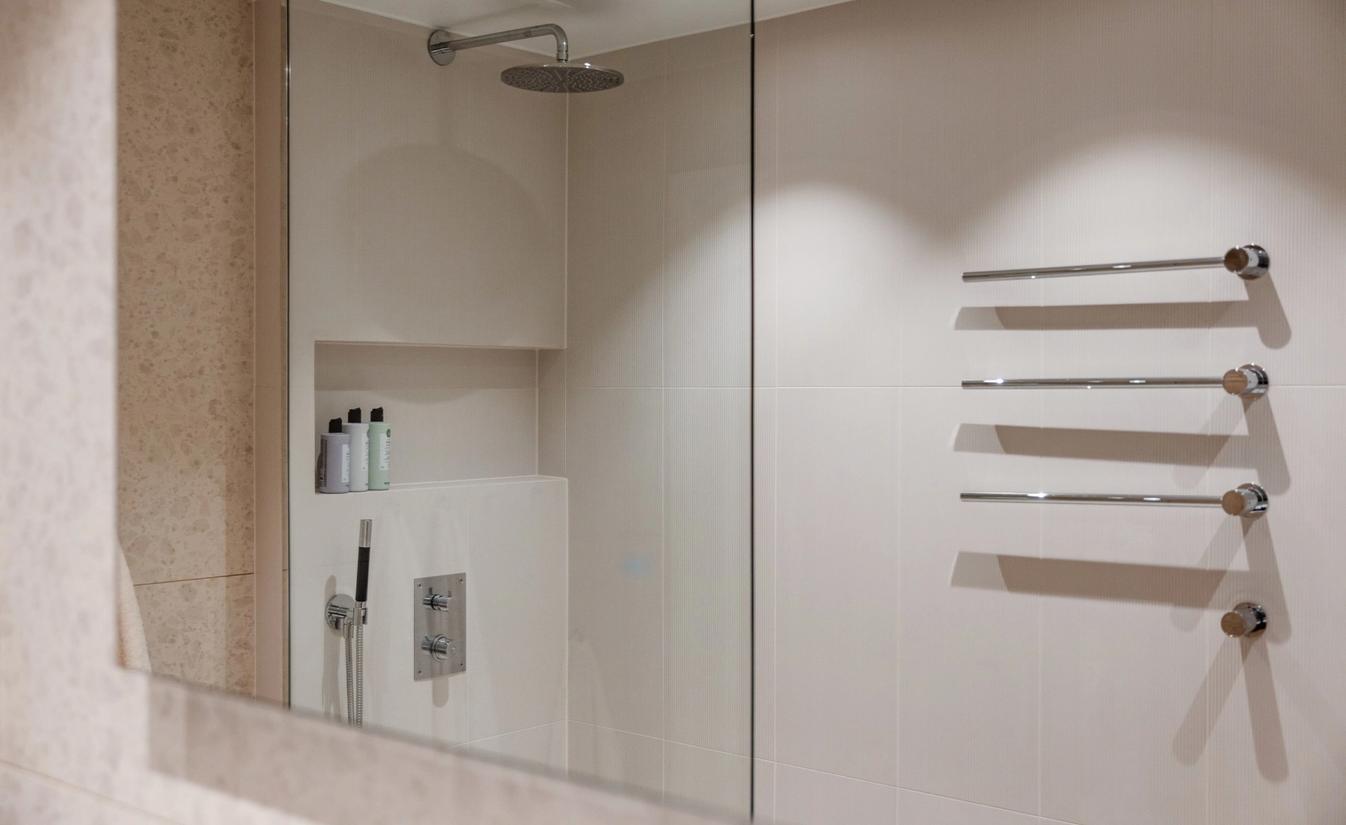 Detaljbilde av lyst bad med store beige fliser på vegg, innebygde armaturer i dusj med egen hyllenisje. Badet er pusset opp av Lomundal.