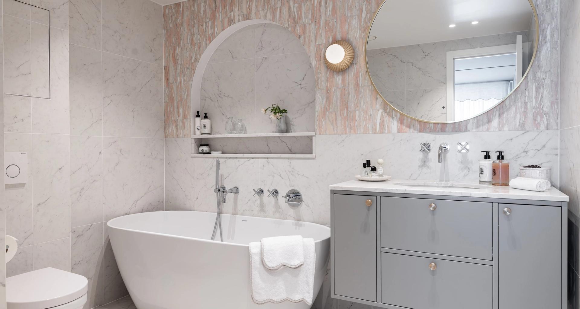 Eksklusivt, klassisk bad i lyse, delikate fargetoner. Badet er flislagt med norsk, rosa Fauske marmor og Carrara marmor. Badet har et frittstående badekar og en buet dusjnisje på vegg. Utført av Lomundal.