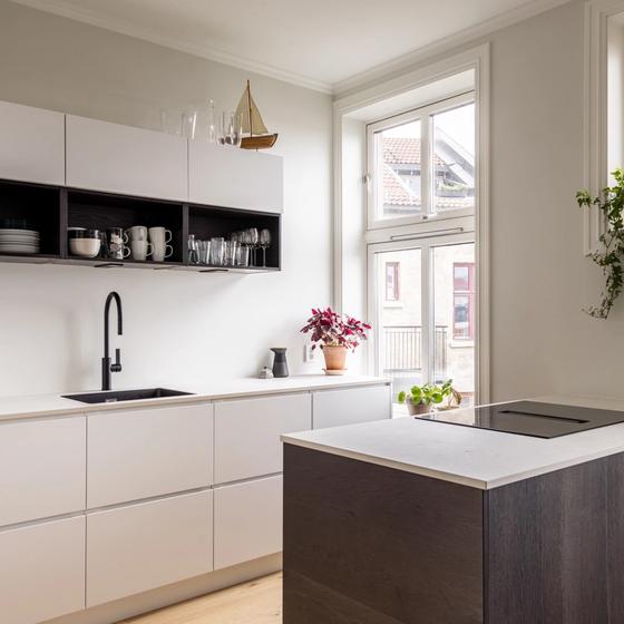 Et moderne kjøkken med lyse og glatte overflater. Sort armatur og kjøkkenøyen i mørkt treverk bryter opp med det lyse og skaper vakre detaljer. God oppbevaringsplass i høyden. Utført av Lomundal.