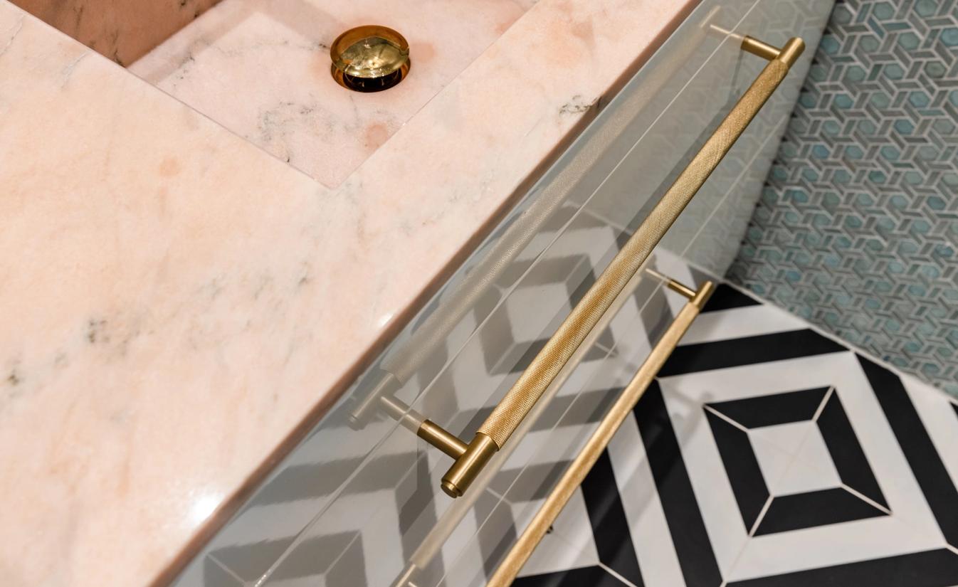 Detaljbilde fra fargerikt bad med blå geometriske fliser på vegg, sorte og hvite geometriske fliser på gulv og baderomsmøbel med rosa benkeplate og detaljer i messing.