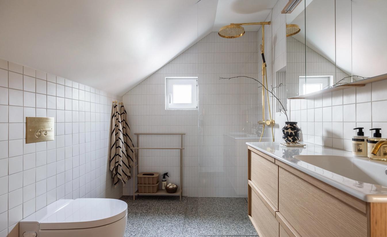 Bad under skråtak med hvite fliser, terrazzogulv, baderomsinnredning i eik og dusj i messing. Utført av Lomundal.