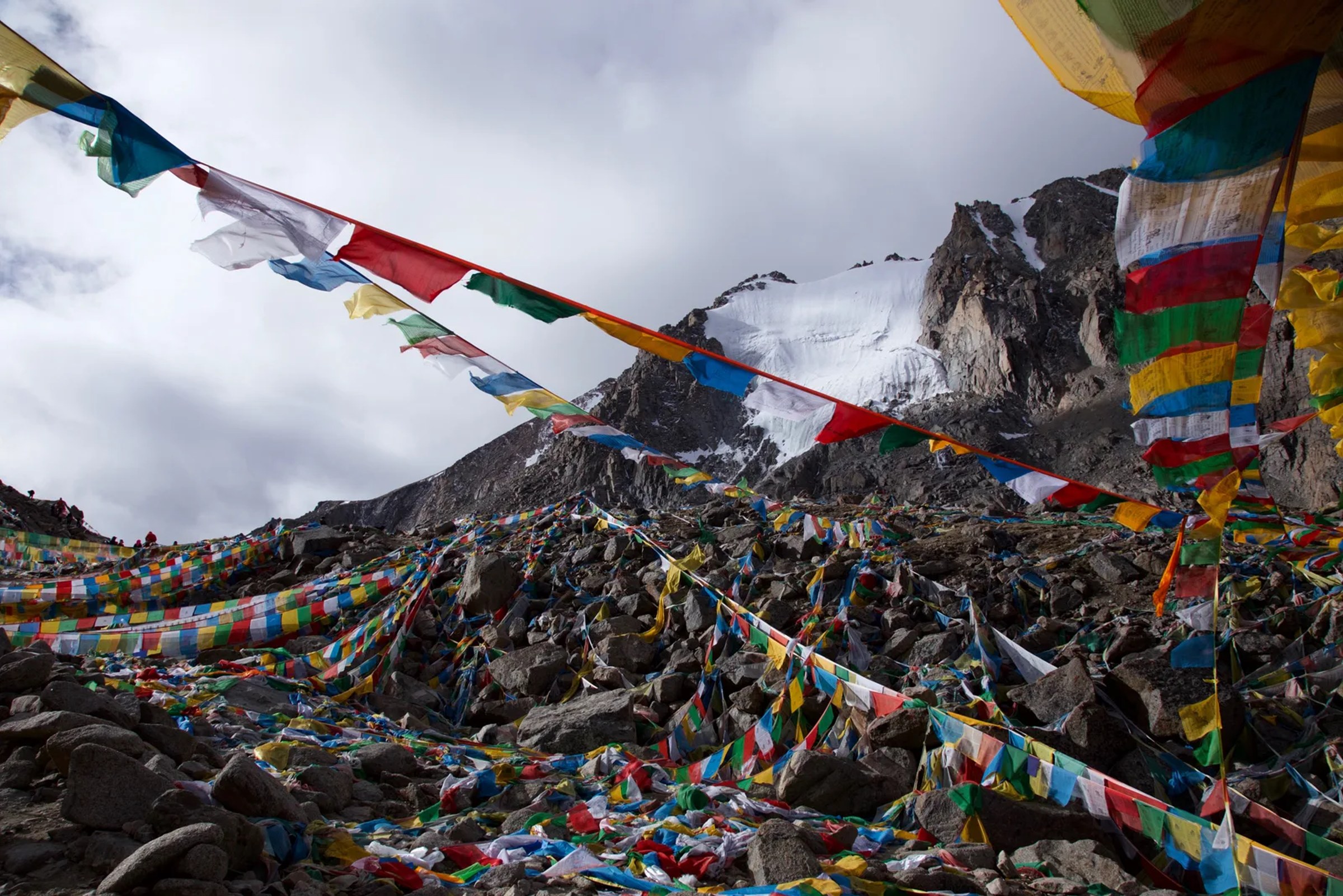 Prayer flags strung across the boulders of Dolma La on the Kailash kora, with a snow-capped rock peak and grey cloud behind