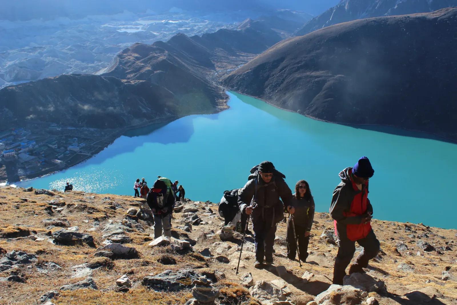 Trekkers working up the rocky Gokyo Ri trail in the early morning, the turquoise second lake and the grey sprawl of the Ngozumpa glacier falling away below them