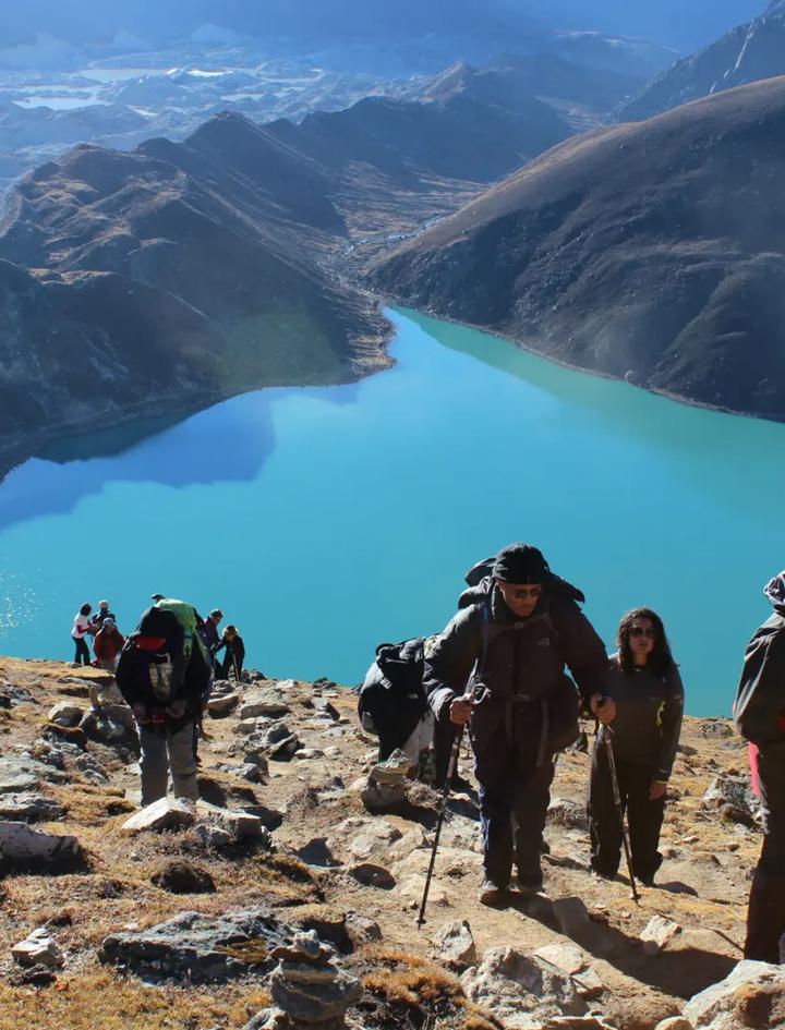 Trekkers working up the rocky Gokyo Ri trail in the early morning, the turquoise second lake and the grey sprawl of the Ngozumpa glacier falling away below them