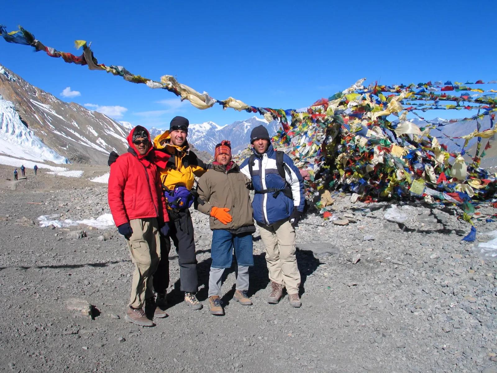Trekkers and their guide in down jackets beside prayer flags on a high Himalayan pass, the kind of cold, exposed 5,000m crossing Larkya La demands
