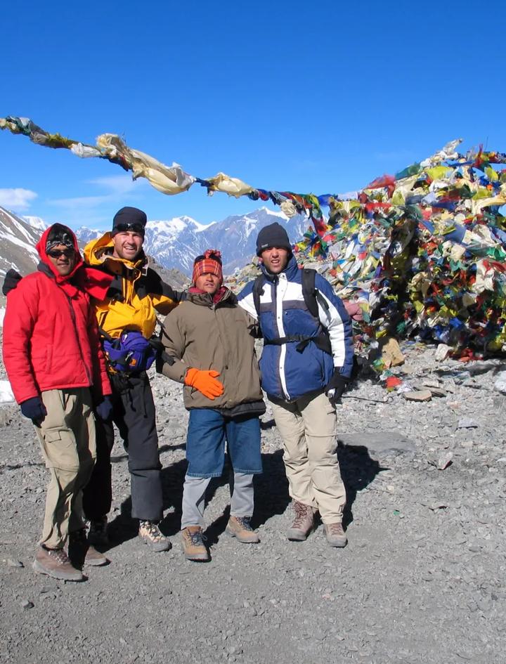 Trekkers and their guide in down jackets beside prayer flags on a high Himalayan pass, the kind of cold, exposed 5,000m crossing Larkya La demands