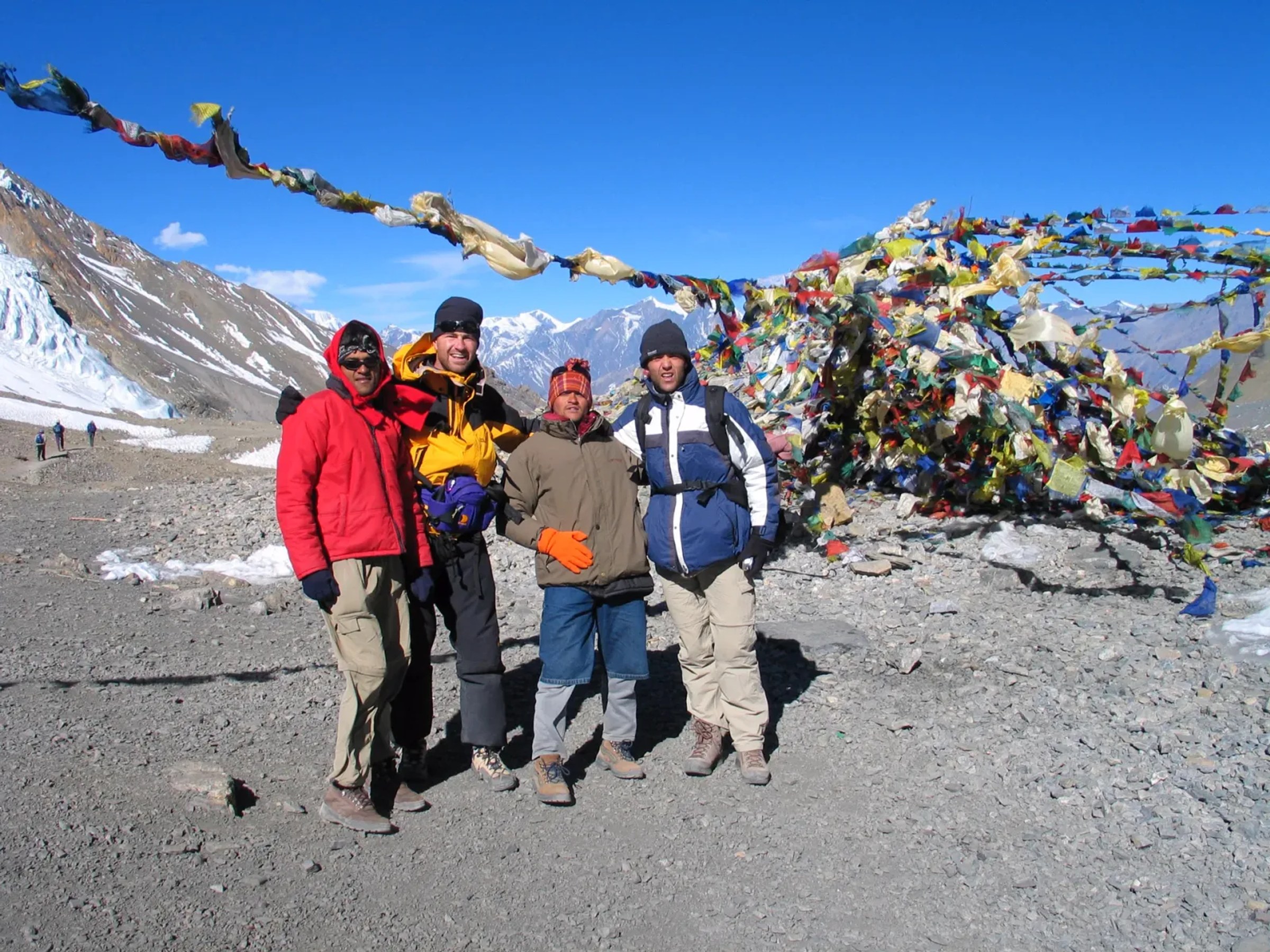 Trekkers and their guide in down jackets beside prayer flags on a high Himalayan pass, the kind of cold, exposed 5,000m crossing Larkya La demands