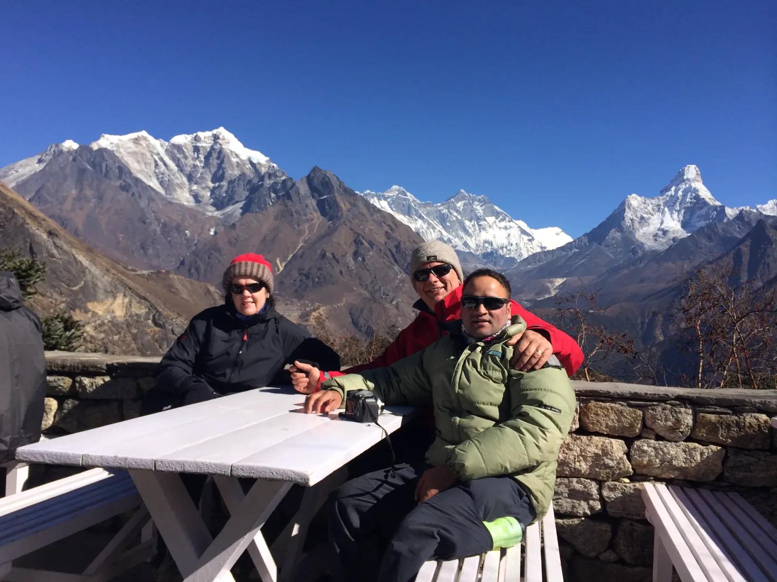 Zwei Gäste und ihr Guide an einem Lodge-Tisch oberhalb von Namche, im Hintergrund Everest, Lhotse und Ama Dablam bei klarem Wetter