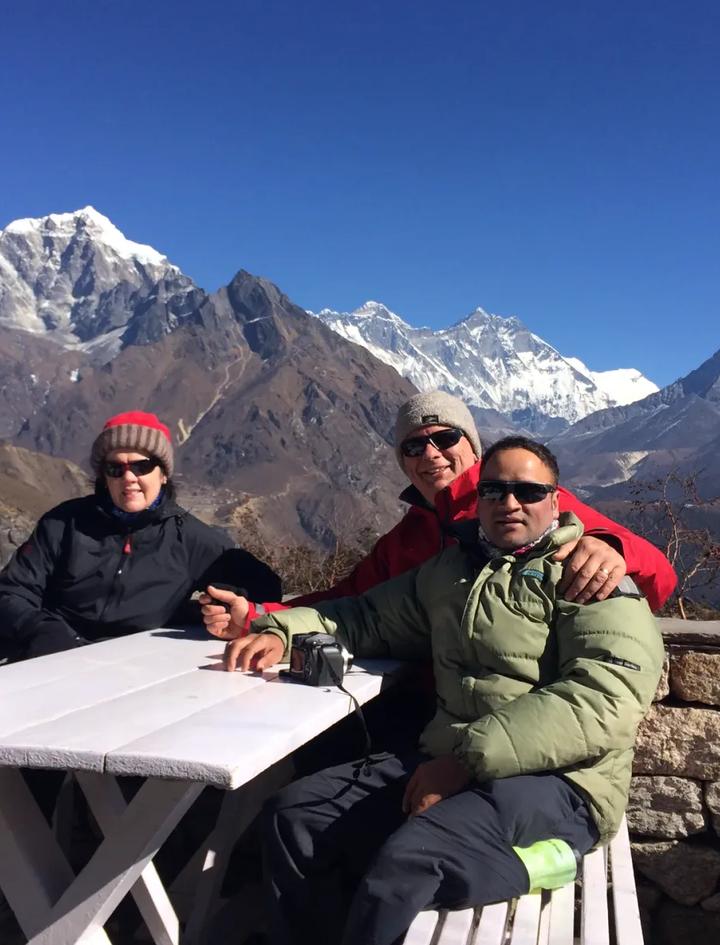 Zwei Gäste und ihr Guide an einem Lodge-Tisch oberhalb von Namche, im Hintergrund Everest, Lhotse und Ama Dablam bei klarem Wetter