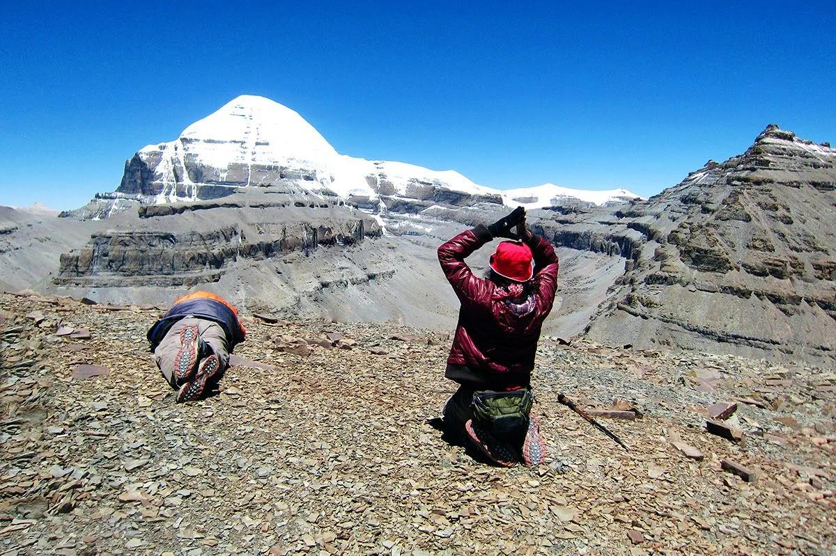 Two pilgrims prostrating on the rocky trail before the snow-covered south face of Mount Kailash under a deep blue sky