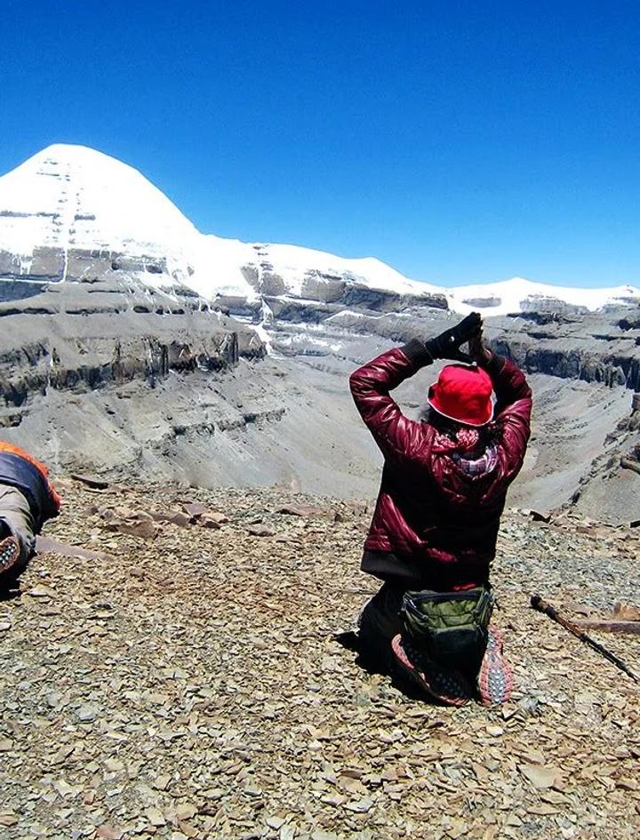 Two pilgrims prostrating on the rocky trail before the snow-covered south face of Mount Kailash under a deep blue sky