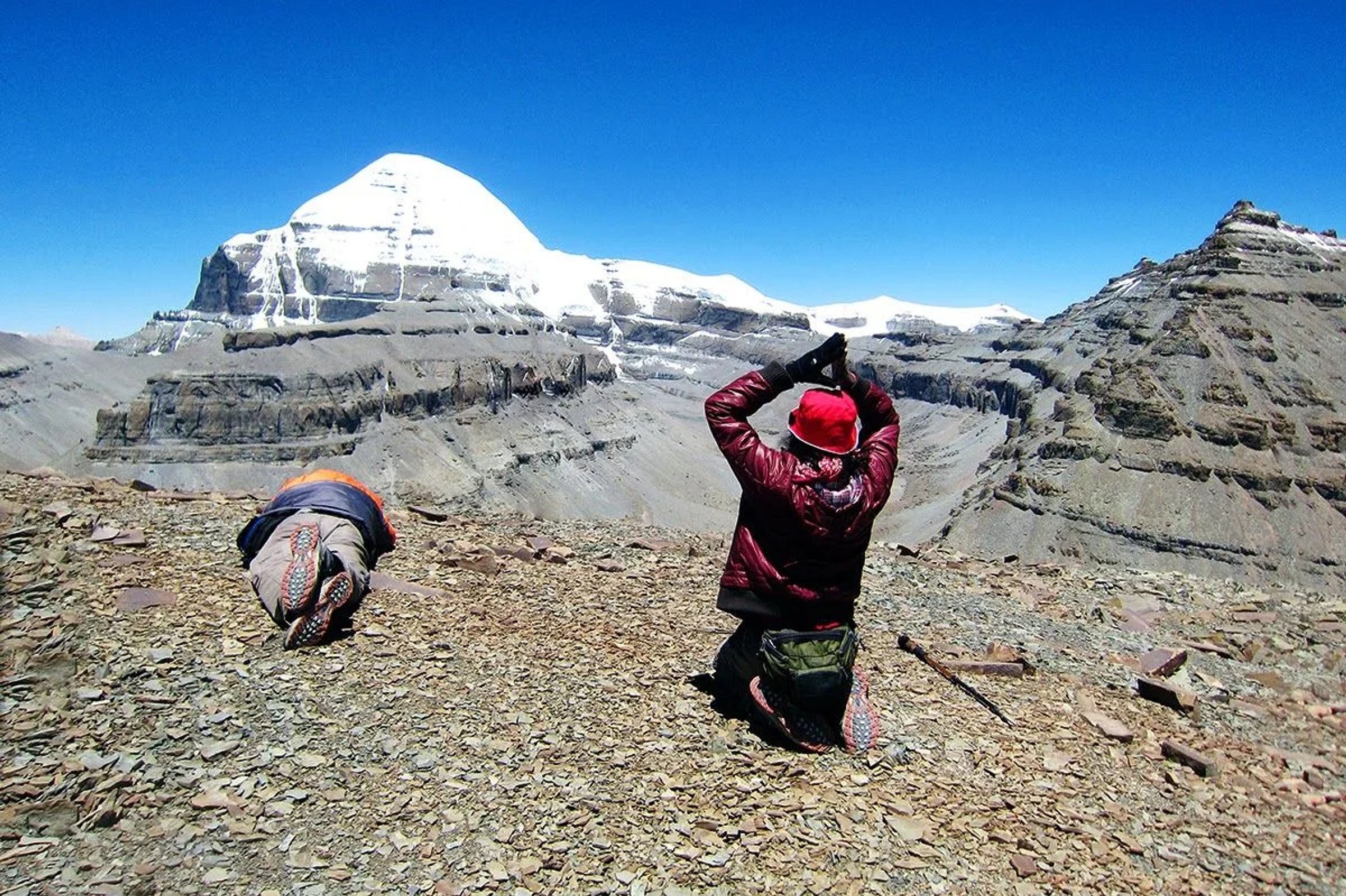 Two pilgrims prostrating on the rocky trail before the snow-covered south face of Mount Kailash under a deep blue sky