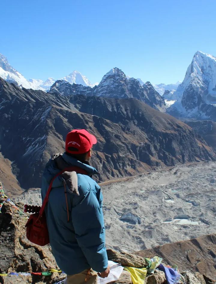 A climber in a down jacket at a prayer-flag cairn above the Ngozumpa glacier, looking across the high Khumbu skyline toward Everest, the same altitude world a Mera Peak expedition climbs through
