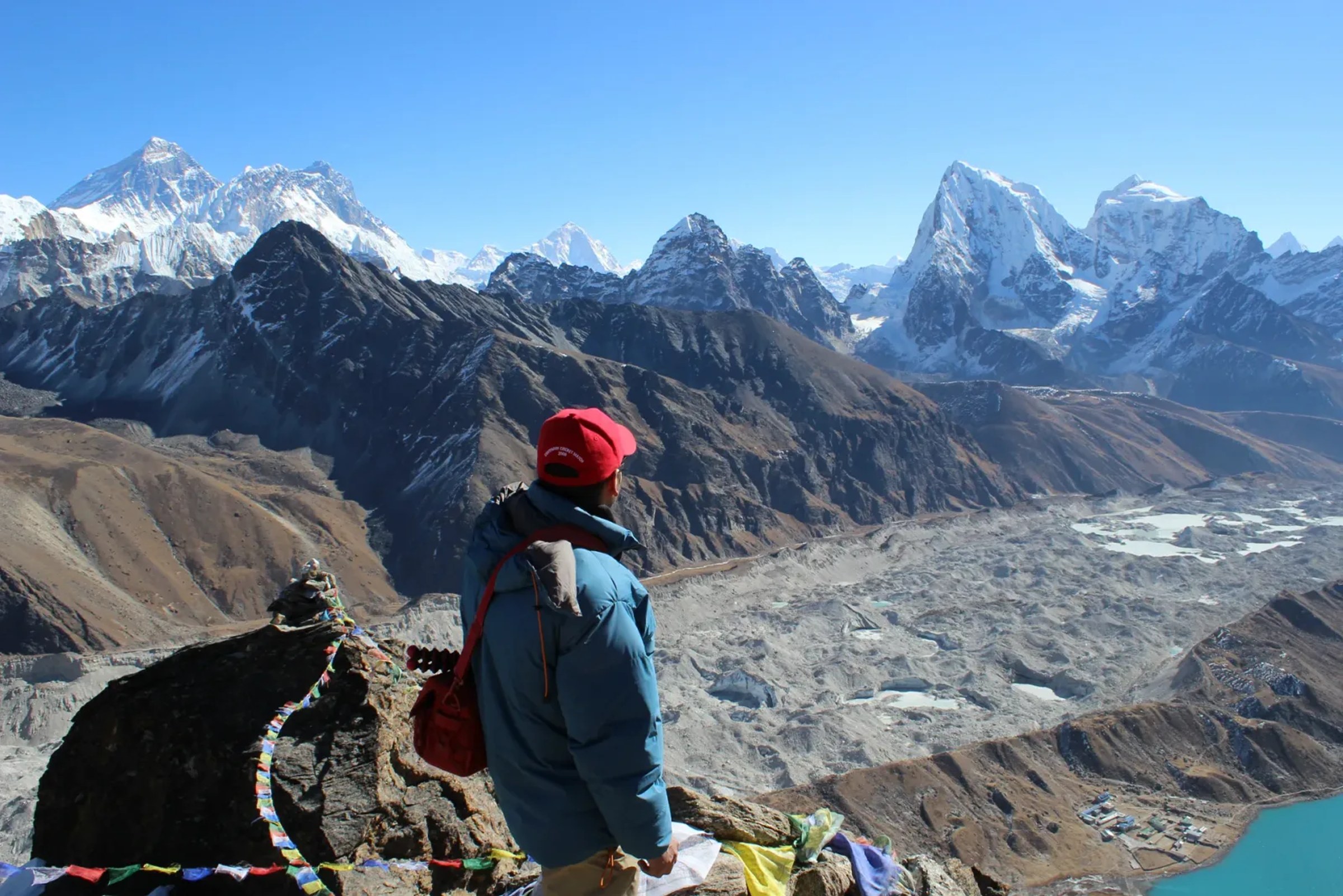A climber in a down jacket at a prayer-flag cairn above the Ngozumpa glacier, looking across the high Khumbu skyline toward Everest, the same altitude world a Mera Peak expedition climbs through
