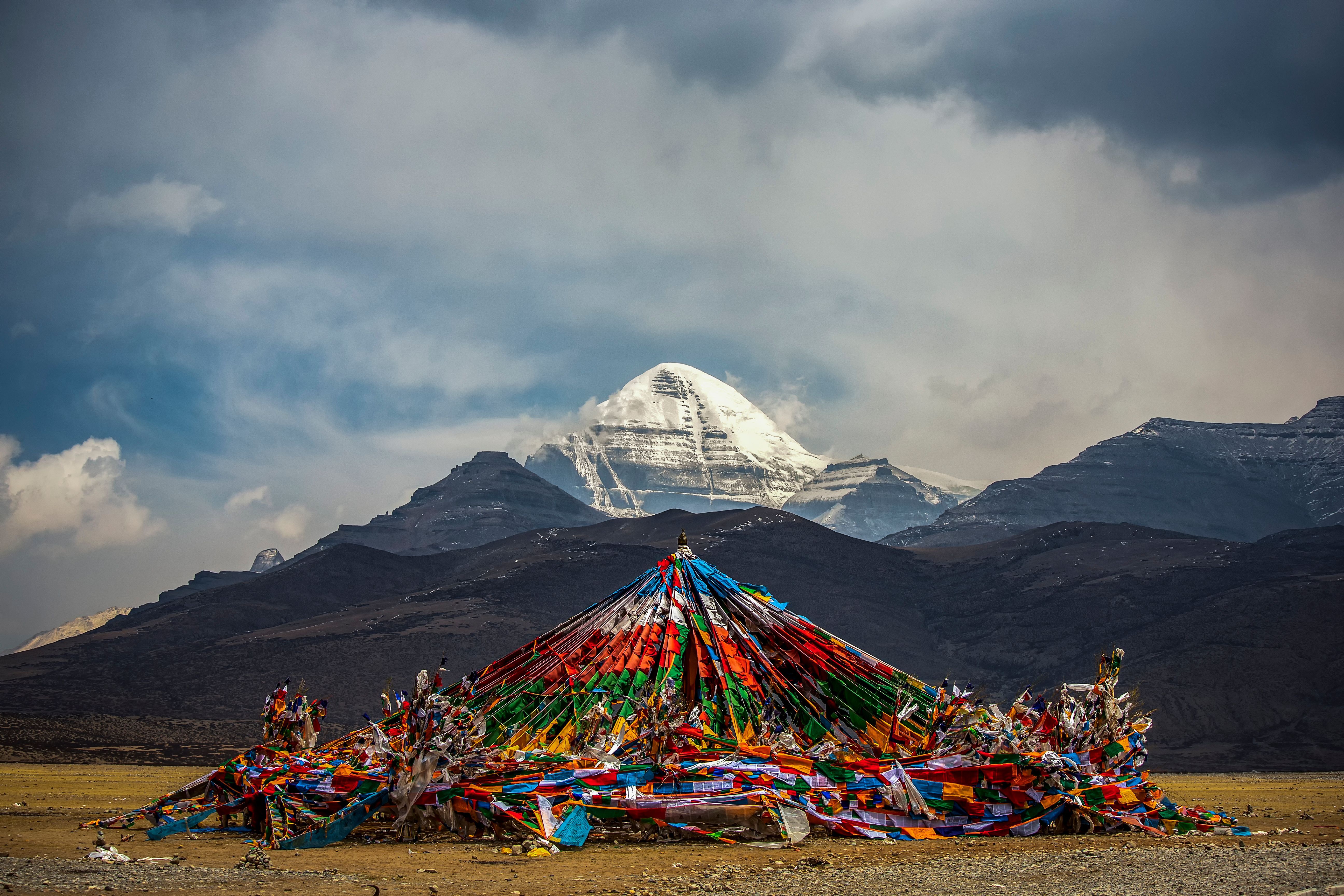 A mound of coloured prayer flags on the open plateau before the snow-covered pyramid of Mount Kailash under a heavy sky