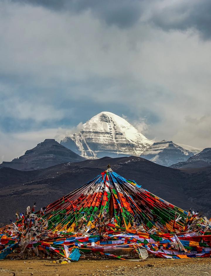 A mound of coloured prayer flags on the open plateau before the snow-covered pyramid of Mount Kailash under a heavy sky