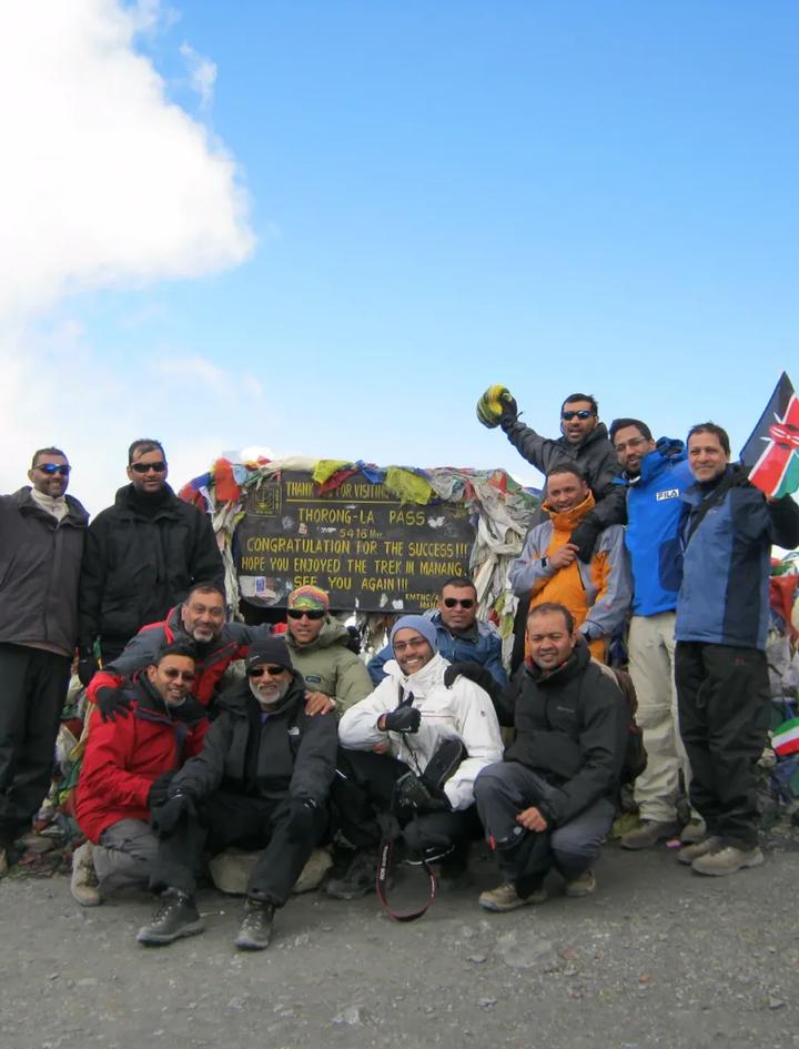 Trekkinggruppe feiert am Schild des Thorong La Pass auf 5.416 Metern, zwischen Bergen aus Gebetsfahnen, Schnee und tiefblauem Himmel