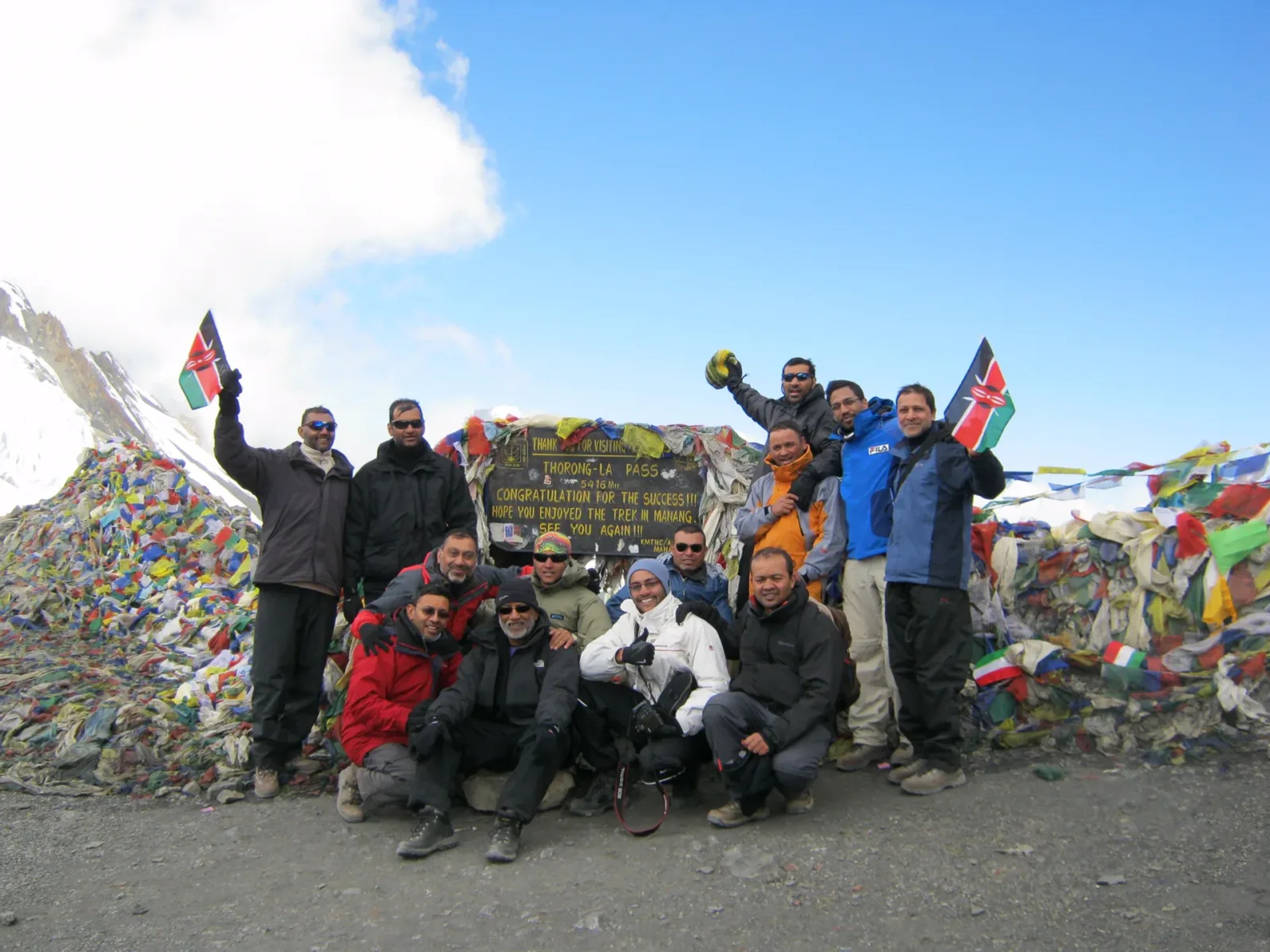 Trekkinggruppe feiert am Schild des Thorong La Pass auf 5.416 Metern, zwischen Bergen aus Gebetsfahnen, Schnee und tiefblauem Himmel