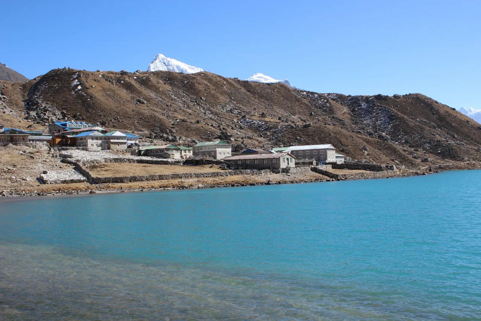 Steinerne Teehäuser von Gokyo am Ufer des türkisfarbenen dritten Gokyo-Sees, dahinter braune Moränenhänge und ein schneebedeckter Gipfel unter klarem Himmel