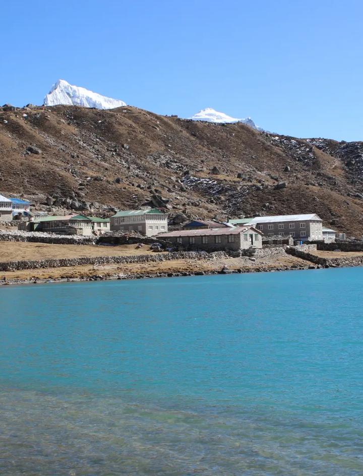 Steinerne Teehäuser von Gokyo am Ufer des türkisfarbenen dritten Gokyo-Sees, dahinter braune Moränenhänge und ein schneebedeckter Gipfel unter klarem Himmel