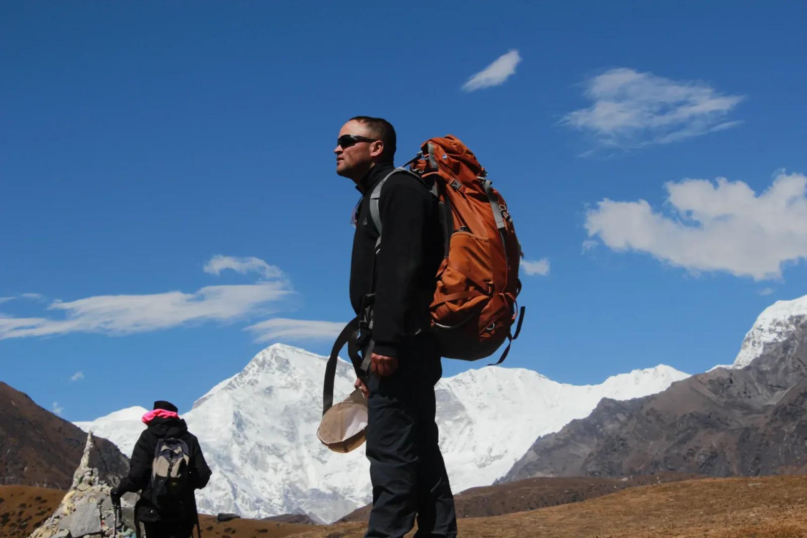 A trekker in profile carrying a large orange pack on a high open trail, silhouetted against deep blue sky with snow-covered peaks and a stone chorten behind