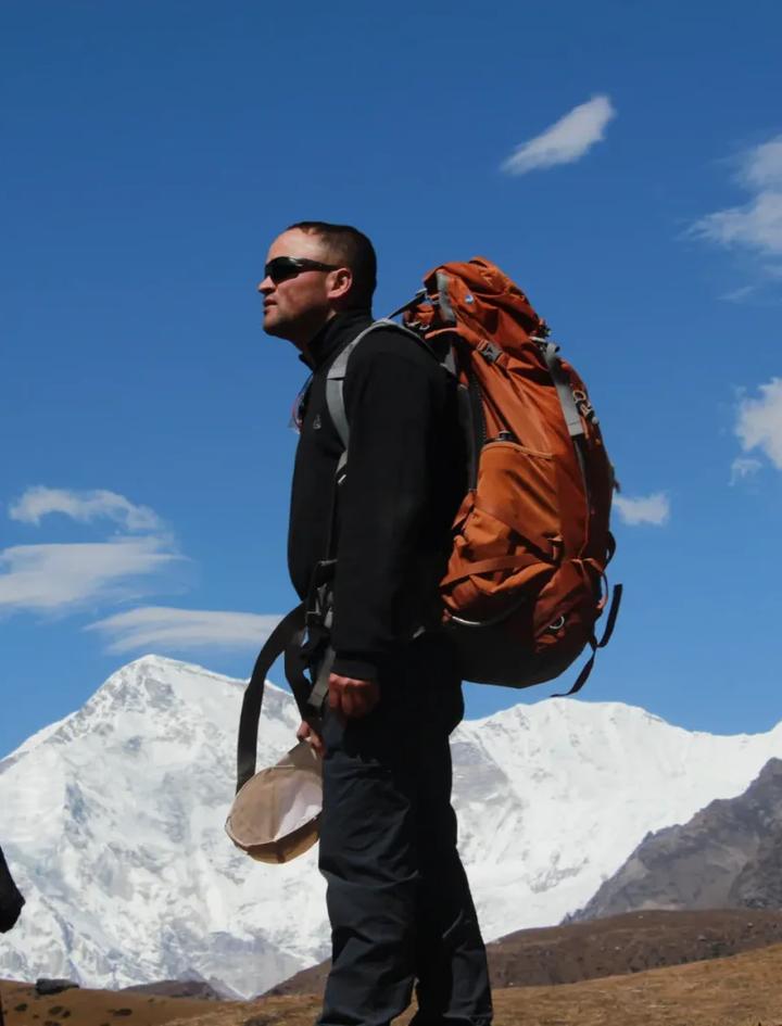 A trekker in profile carrying a large orange pack on a high open trail, silhouetted against deep blue sky with snow-covered peaks and a stone chorten behind