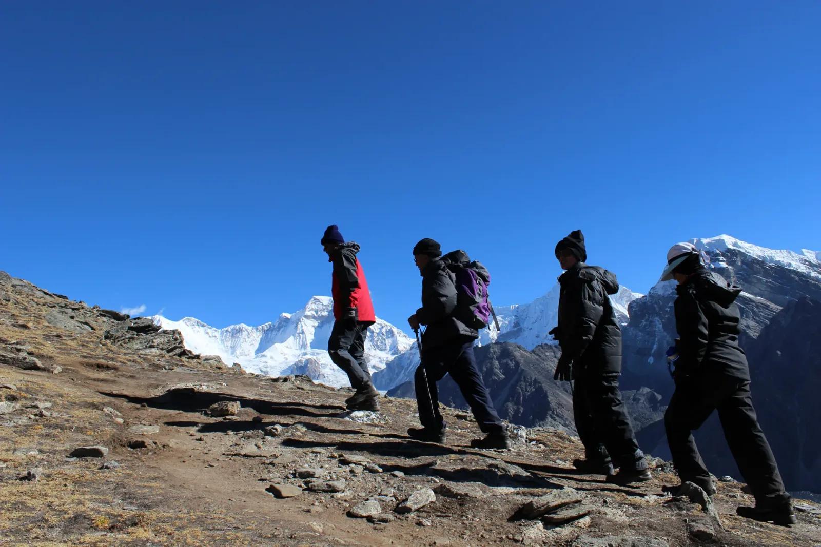 Four trekkers in down jackets and wool hats walking single file along a rocky ridgeline against a deep blue sky, with snow-covered Himalayan peaks filling the horizon behind them