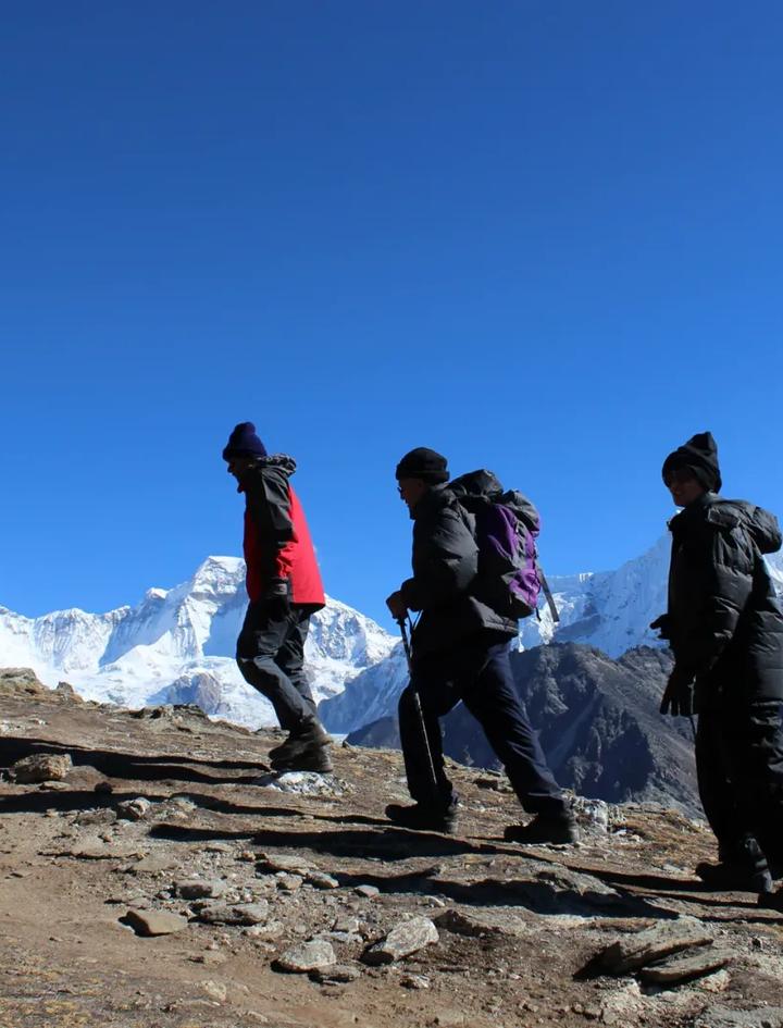 Four trekkers in down jackets and wool hats walking single file along a rocky ridgeline against a deep blue sky, with snow-covered Himalayan peaks filling the horizon behind them