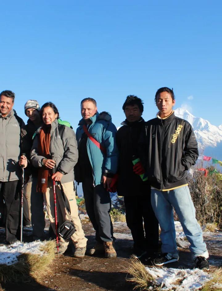 Eine Trekkerin mit ihrer Gruppe und einheimischen Guides am Aussichtspunkt des Poon Hill im Morgenlicht, hinter ihnen schneebedeckte Himalaya-Gipfel und Gebetsfahnen