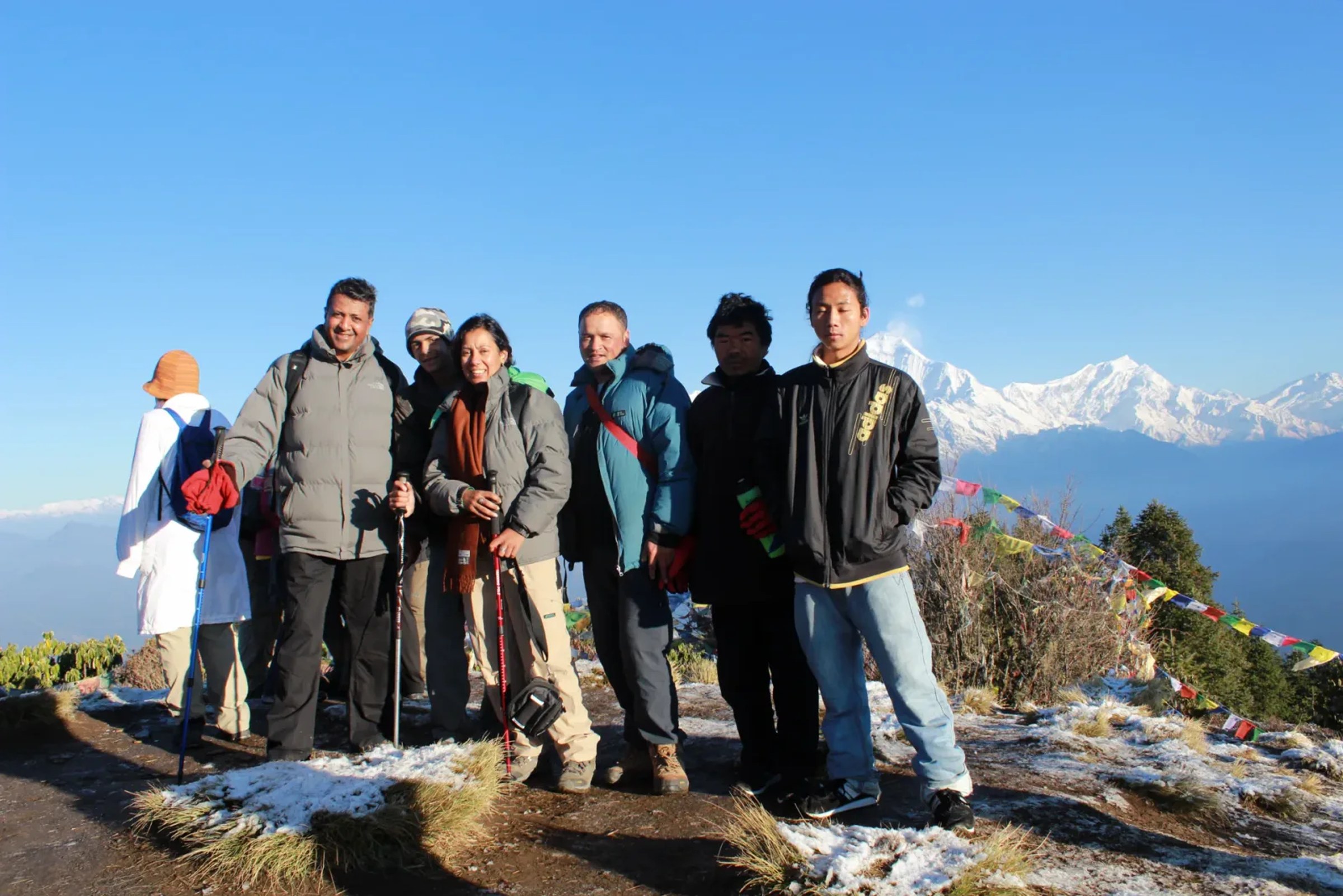 Eine Trekkerin mit ihrer Gruppe und einheimischen Guides am Aussichtspunkt des Poon Hill im Morgenlicht, hinter ihnen schneebedeckte Himalaya-Gipfel und Gebetsfahnen