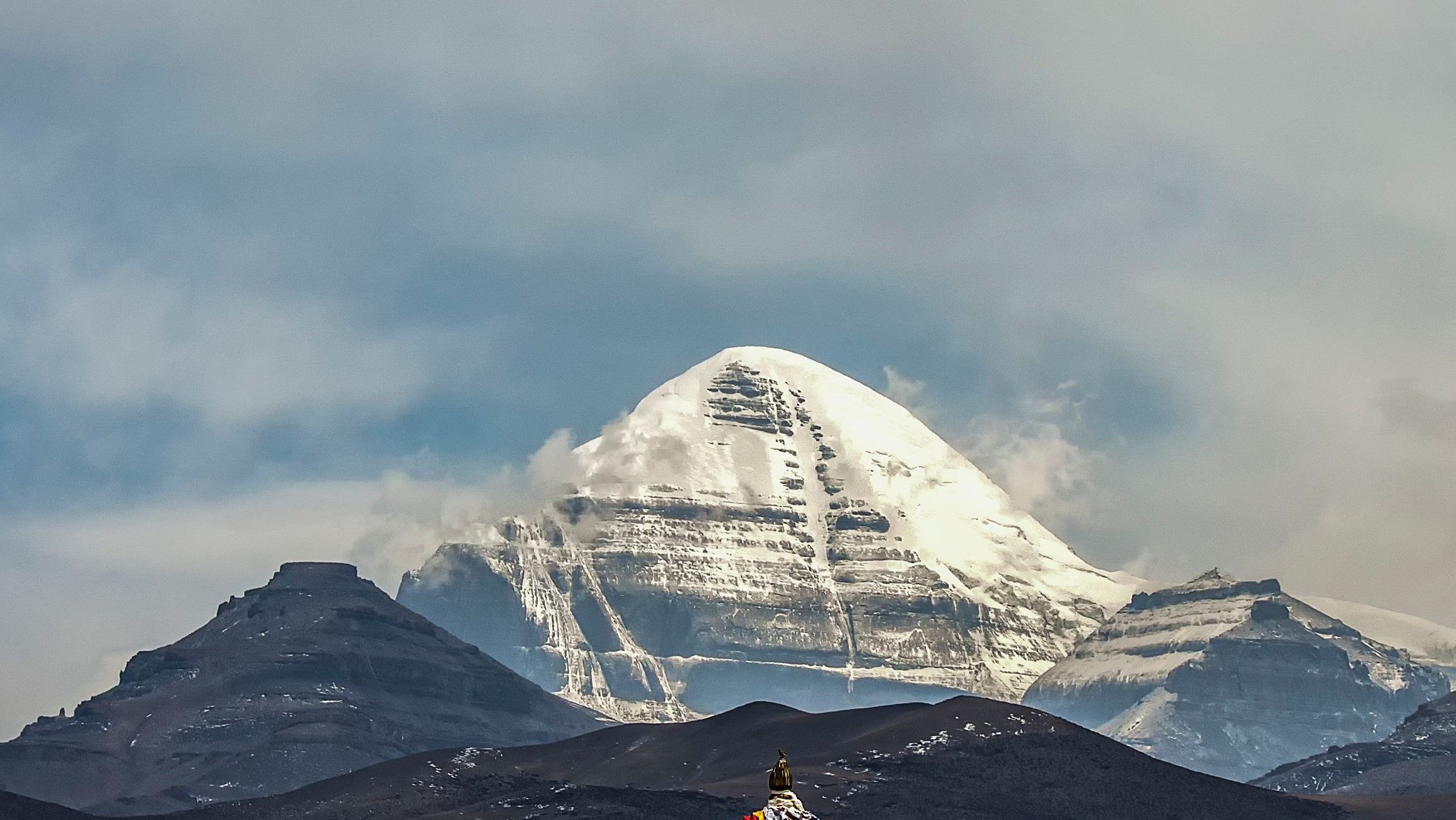 The snow-striped south face of Mount Kailash rising behind a dark foreground ridge under storm cloud, with the tip of a prayer-flag mound just visible below
