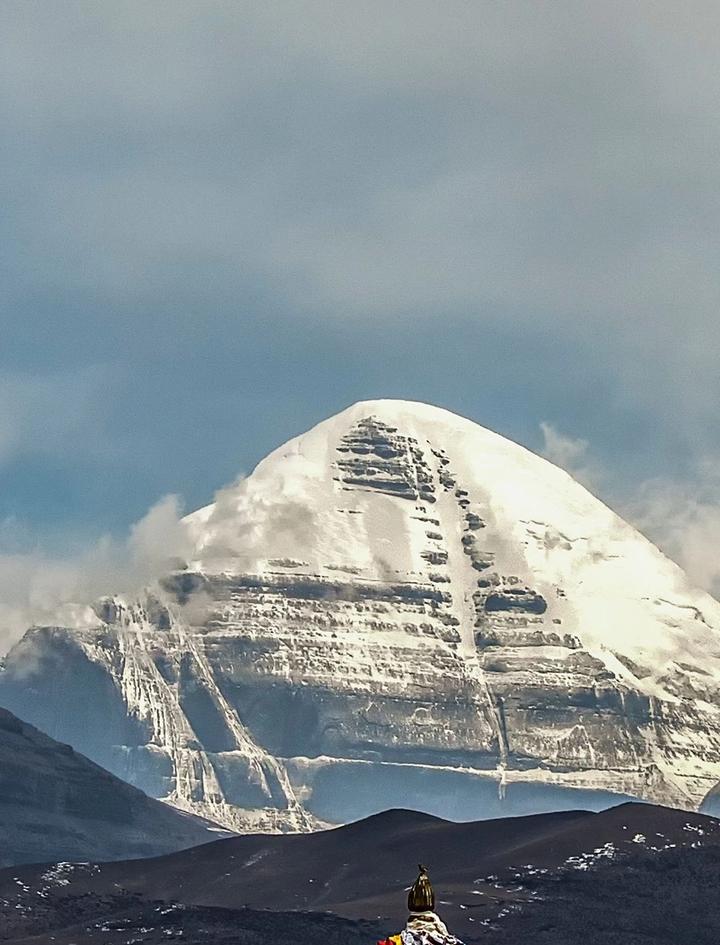 The snow-striped south face of Mount Kailash rising behind a dark foreground ridge under storm cloud, with the tip of a prayer-flag mound just visible below
