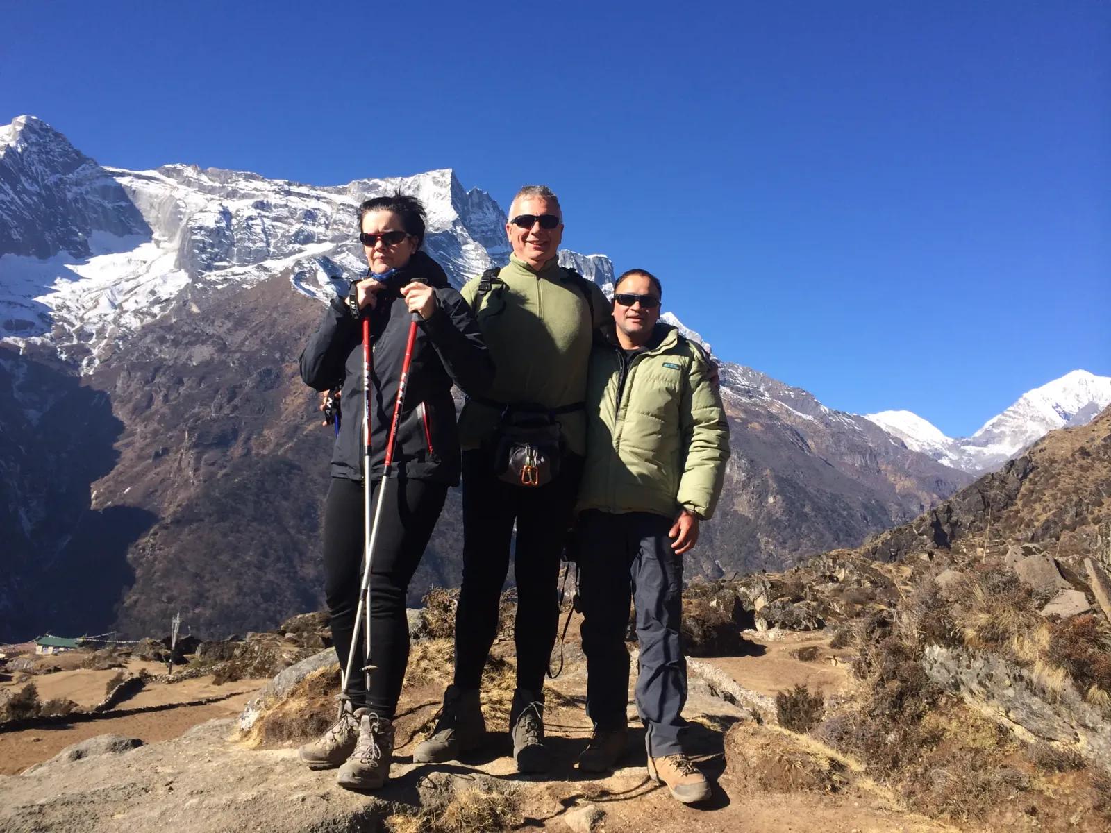 Ein einheimischer Trekkingguide mit zwei Gästen auf einem Aussichtspunkt hoch über dem Khumbu-Tal, hinter ihnen schneebedeckte Sechstausender unter tiefblauem Himmel