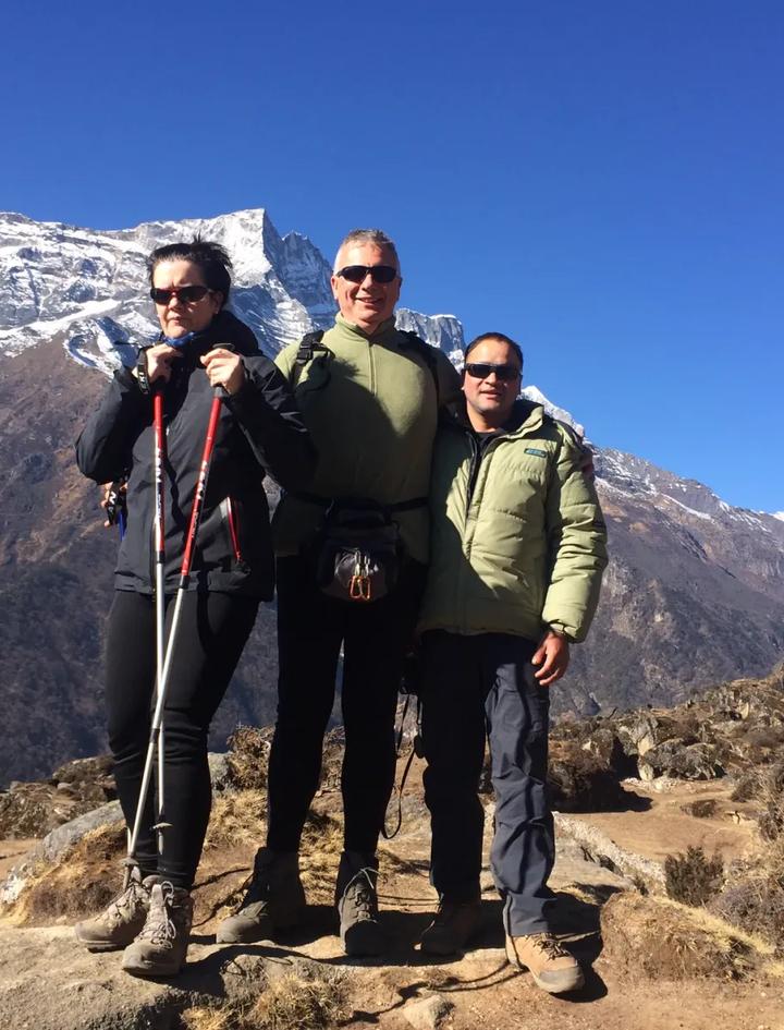 Ein einheimischer Trekkingguide mit zwei Gästen auf einem Aussichtspunkt hoch über dem Khumbu-Tal, hinter ihnen schneebedeckte Sechstausender unter tiefblauem Himmel