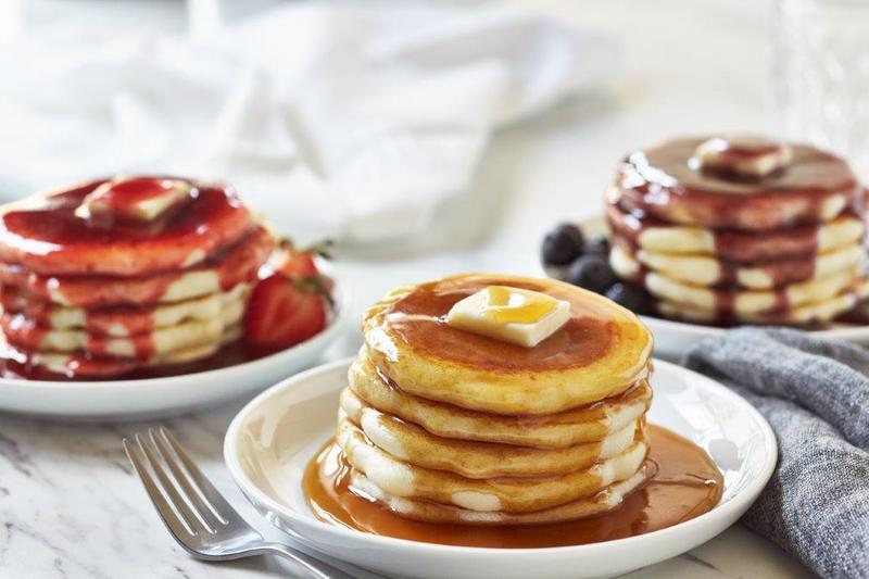 Drie stapels pancakes op witte borden: met siroop en boter, met aardbeiensaus en met bosbessensaus