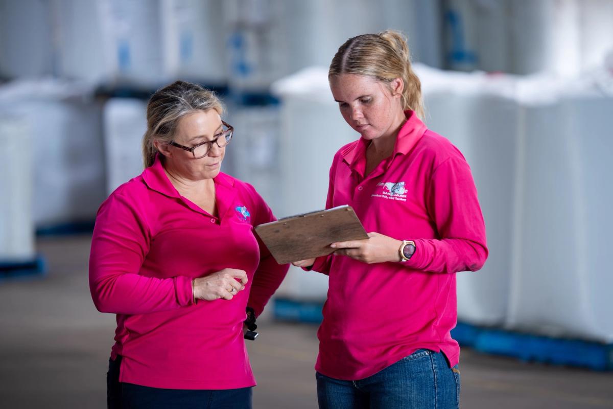 Two Humpty Doo Barramundi staff in pink polo shirts reviewing a clipboard in a production facility with IBC tanks in the background.