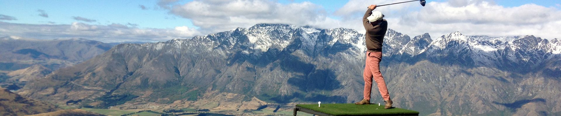 A golfer teeing off above Queenstown with The Remarkables behind them