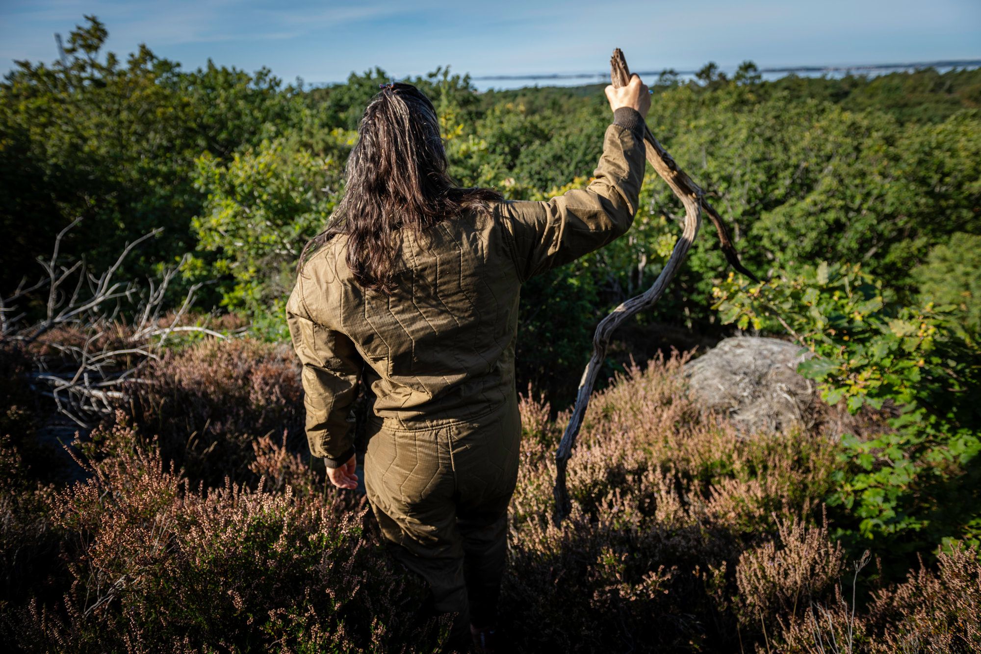 Stine står i ljung och blåbärsris med en stor gren lyft mot himlen.