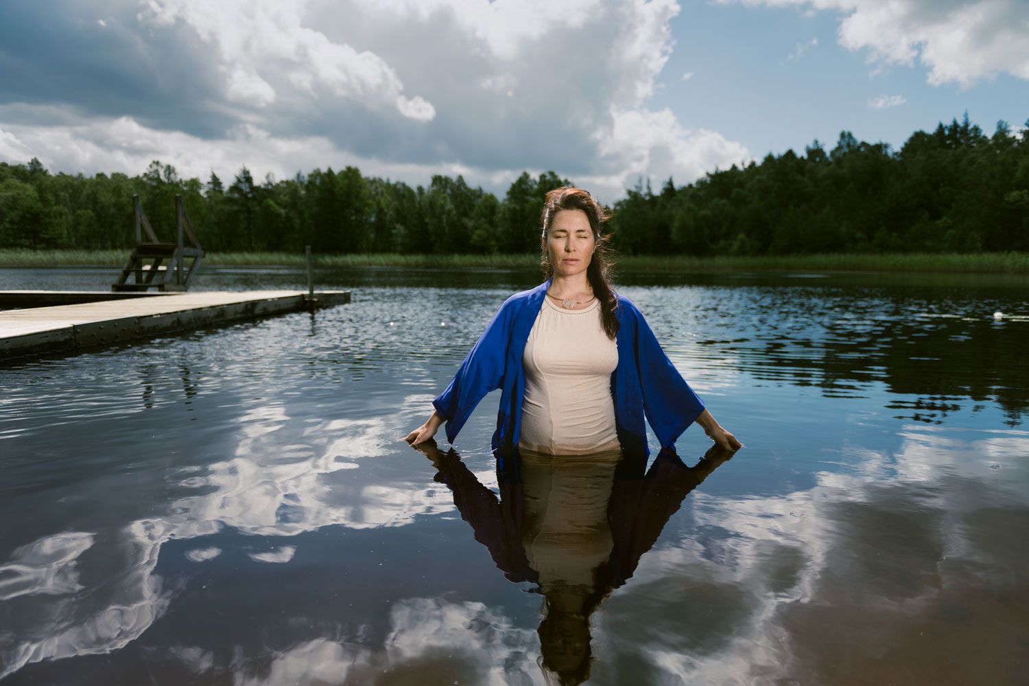 Stine står i vatten upp till knäna i en stilla sjö, med skog och en brygga bakom sig.