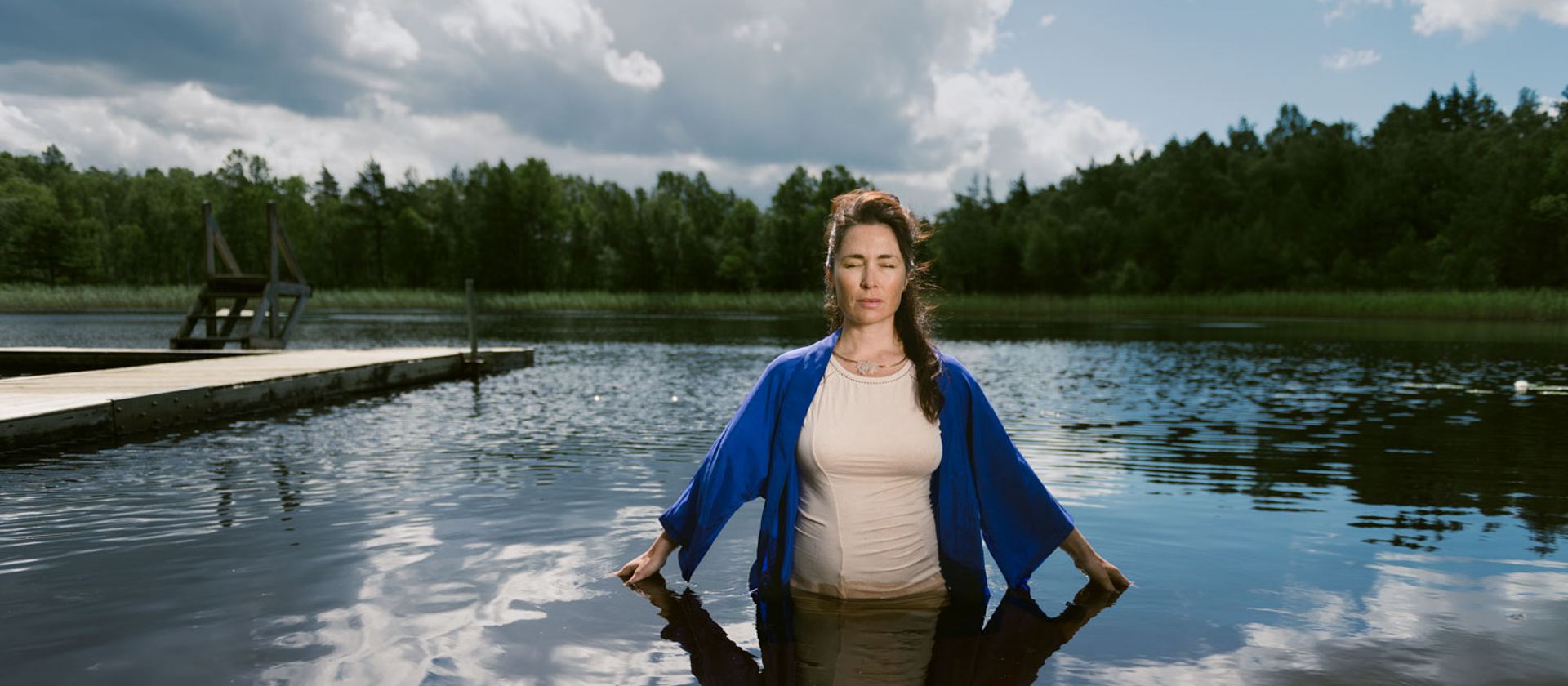 Stine står i vatten upp till knäna i en stilla sjö, med skog och en brygga bakom sig.