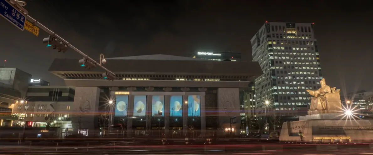 A nighttime view of a building with ornate pillars and detailed images on its facade, observed from the opposite street side.