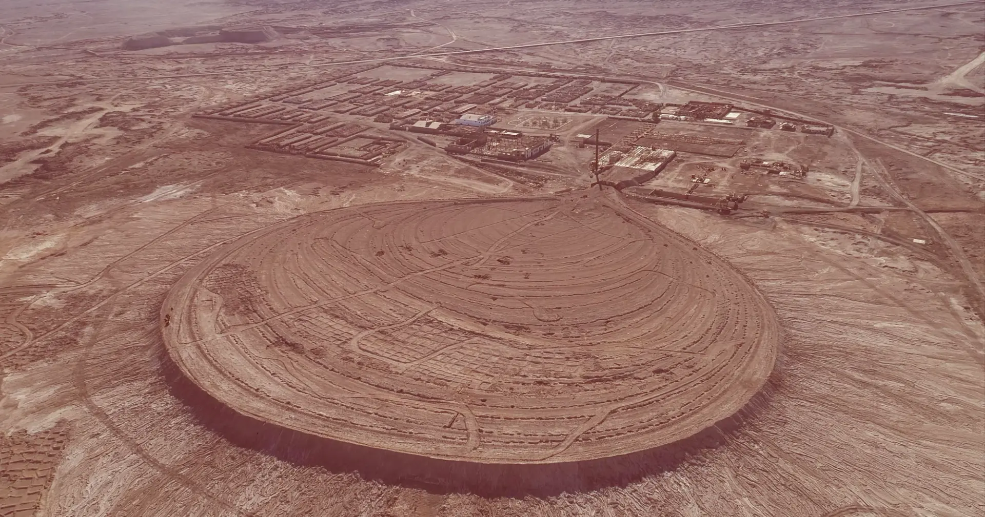 Overhead view of a landscape featuring a large circular earthen structure in the center, surrounded by various buildings a...