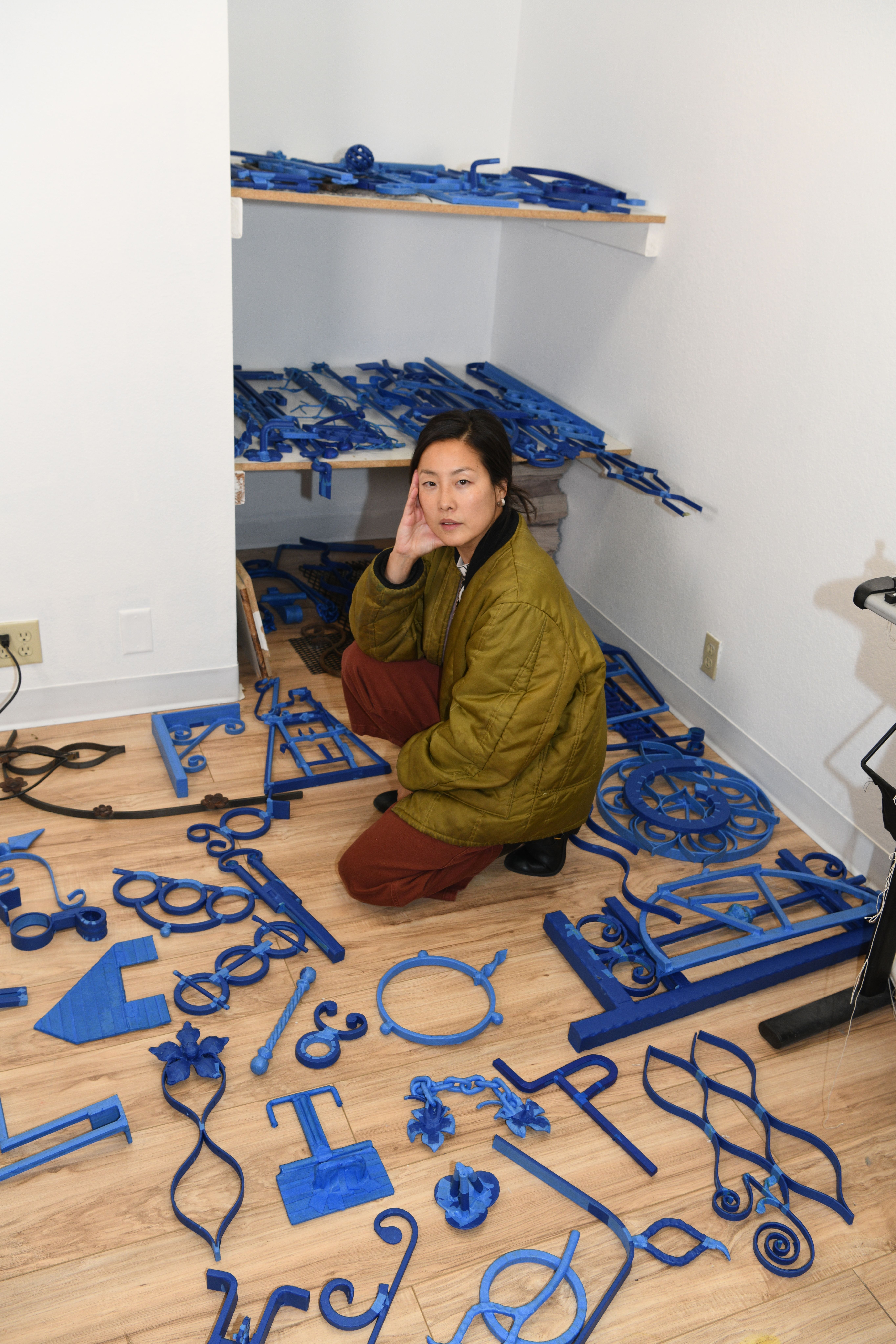 Artist sitting thoughtfully among scattered blue sculptural elements on a studio floor, reflecting a creative process in motion.