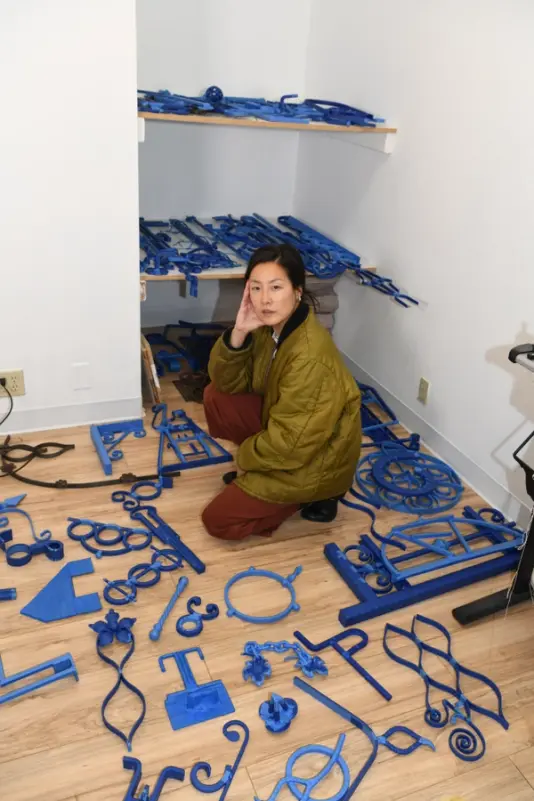 Artist sitting thoughtfully among scattered blue sculptural elements on a studio floor, reflecting a creative process in motion.